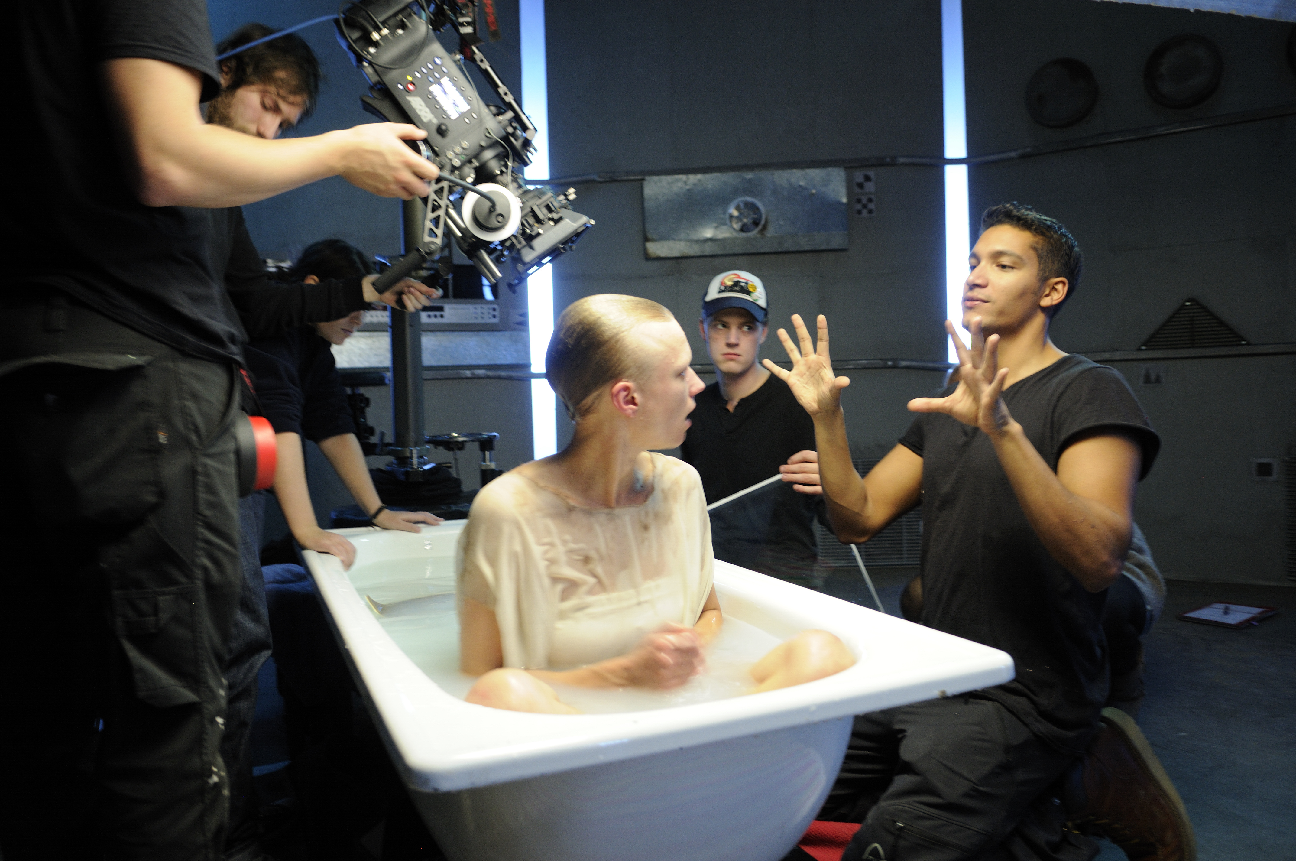 Filmcrew rundt et badekar fylt med hvit væske hvor det sitter en hvitkledd skuespiller som blir instruert av regissør.